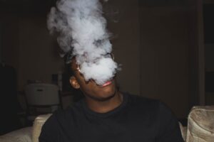Young man exhaling a large cloud of vapor smoke, obscuring his face, wearing glasses and a black t-shirt indoors.