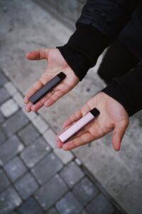 Two hands holding gray and pink pod vapes outdoors on a cobblestone surface, wearing a black jacket.